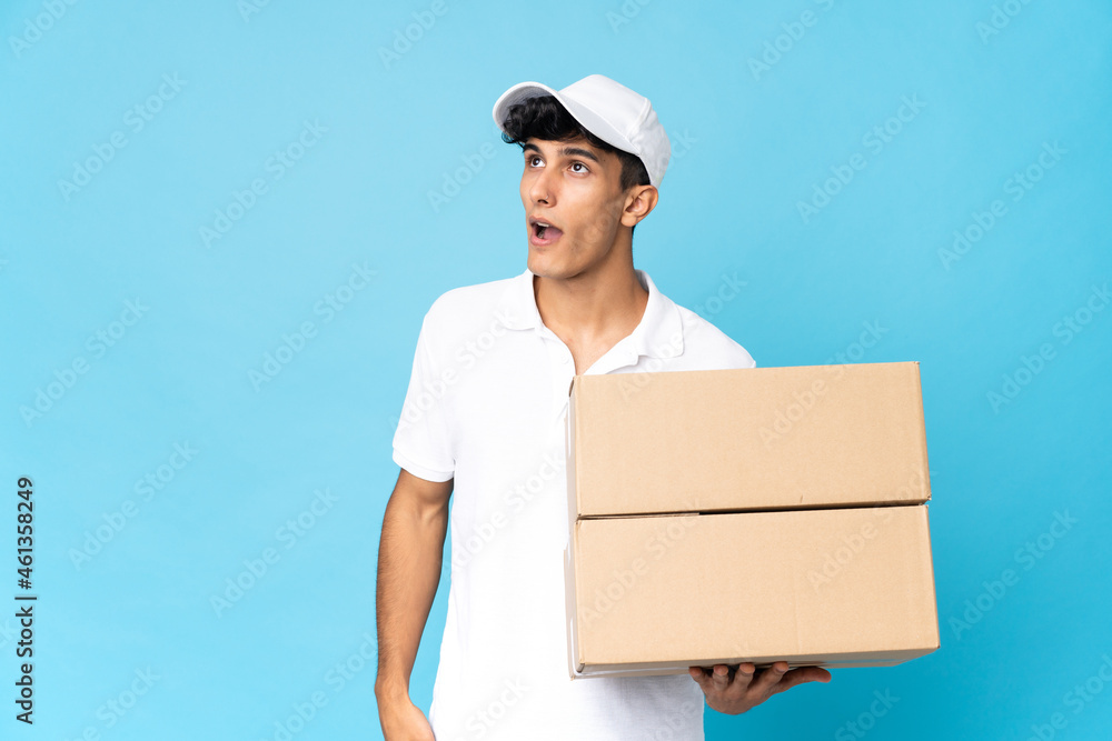 Delivery Argentinian man isolated on blue background looking up and with surprised expression Delivery Argentinian man isolated on blue background looking up and with surprised expression