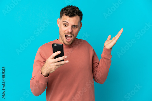 Young caucasian handsome man isolated on blue background looking at the camera while using the mobile with surprised expression