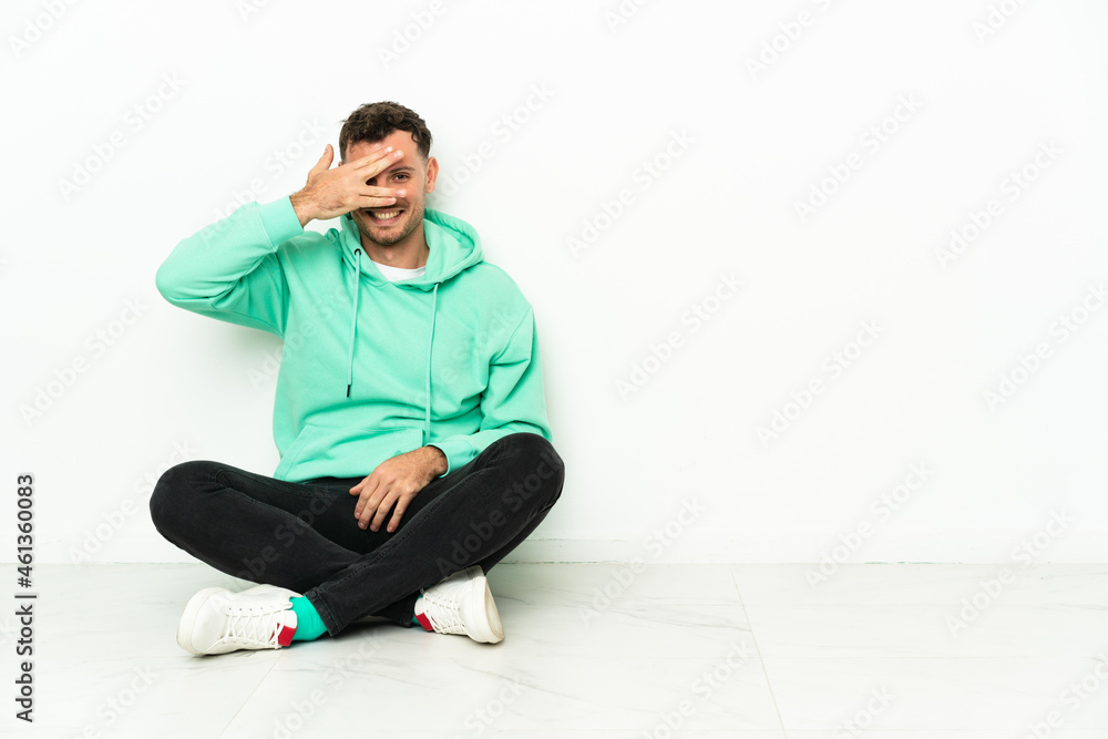 Young handsome caucasian man sitting on the floor covering eyes by hands and smiling Young handsome caucasian man sitting on the floor covering eyes by hands and smiling