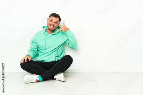 Young handsome caucasian man sitting on the floor making phone gesture. Call me back sign