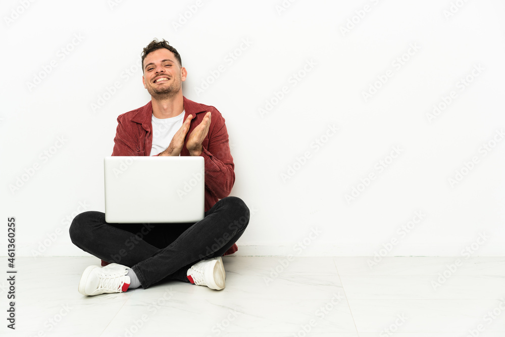 Young handsome caucasian man sit-in on the floor with laptop applauding after presentation in a conference Young handsome caucasian man sit-in on the floor with laptop applauding after presentation in a conference