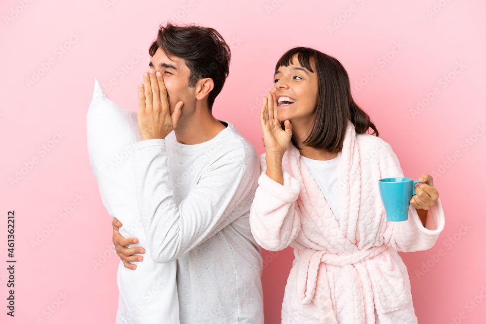 Young couple in pajamas isolated on pink background shouting with mouth wide open to the lateral Young couple in pajamas isolated on pink background shouting with mouth wide open to the lateral