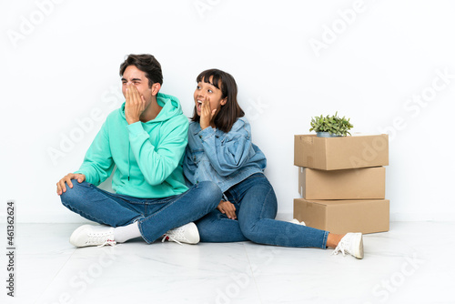 Young couple making a move while picking up a box full of things sitting on the floor isolated on white background shouting with mouth wide open to the lateral © luismolinero