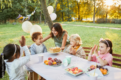Portrait of smiling mother bringing Birthday cake to son during Birthday party outdoors in sunlight