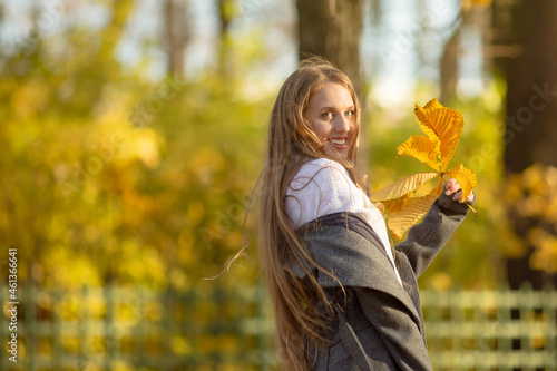 Attractive happy woman holding yellow autumn leaves in her hands. Portrait of a pretty young woman with light brown hair on a background of golden foliage in the park. Joyful autumn mood. Fall season.