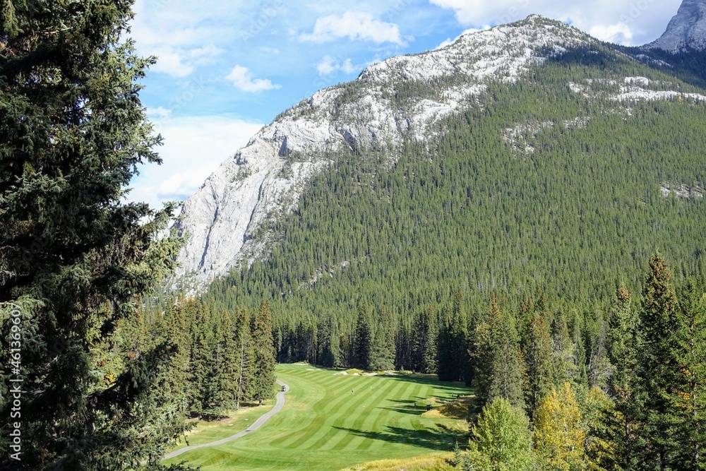 A beautiful view of a par 4 golf hole on a course with a huge mountain in the background, surrounded by forest, on a beautiful sunny day with blue sky, in the rocky mountains near Banff, Alberta