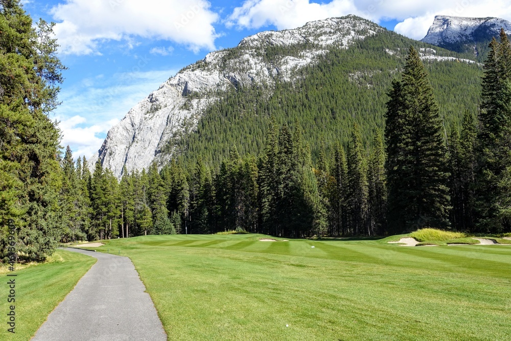 A beautiful view of a par 4 golf hole on a course with a huge mountain in the background, surrounded by forest, on a beautiful sunny day with blue sky, in the rocky mountains near Banff, Alberta