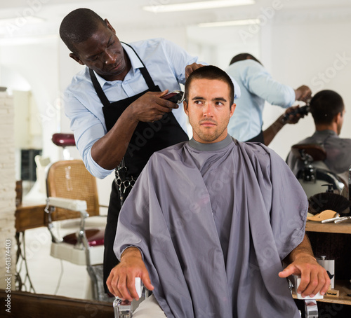 Young man getting haircutting with electric clipper by professional African barber in salon..