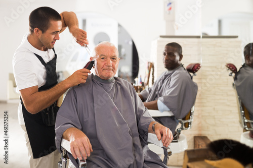 Elderly man getting haircut with electric clipper from skillful young barber in salon..