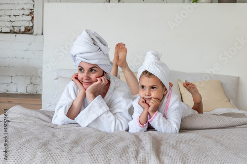 Mom and daughter are dressed in white terry robes, lie on the bed. Nursing communication in the family.