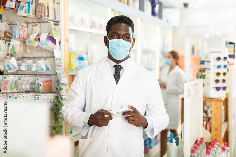 Portrait of African-american male pharmacist in face mask who standing in salesroom of drugstore and holding drug package in hands