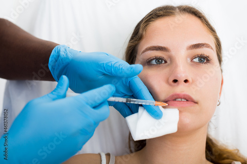 Closeup face of young female client receiving injections during lip enhancement procedure, professional cosmetologist hands in rubber gloves holding syringe..