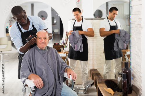 Elderly man getting haircutting with electric clipper from professional African barber in salon