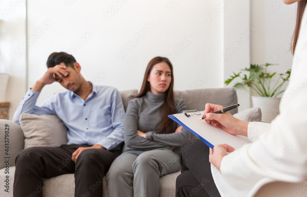 Stressed young Asian male and woman sitting on sofa and talking with professional psychologist while conducting a consultation and making notes Stressed young Asian male and woman sitting on sofa and talking with professional psychologist while conducting a consultation and making notes