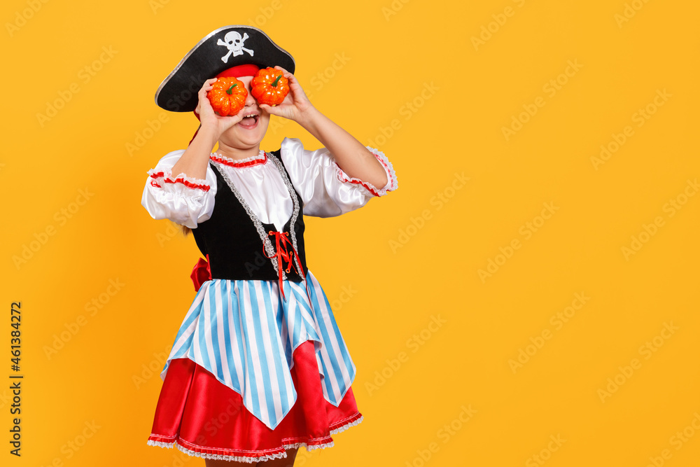Celebrating Halloween. Cheerful girl in a carnival costume of a pirate in the studio on a yellow background. The child holds the pumpkins near his eyes.