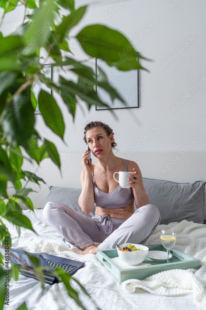 Young woman in comfortable lilac pajamas works on a computer and talks a cell phone while lying the bed in the bedroom. Young woman in comfortable lilac pajamas works on a computer and talks a cell phone while lying the bed in the bedroom.
