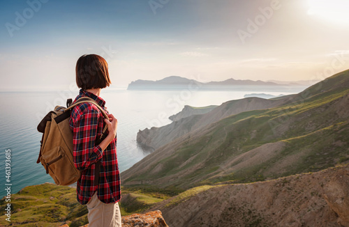 A young Asian woman with a backpack hiking in the summer enjoying a sunny view of the sea. Mountain and coastal travel, freedom and an active lifestyle