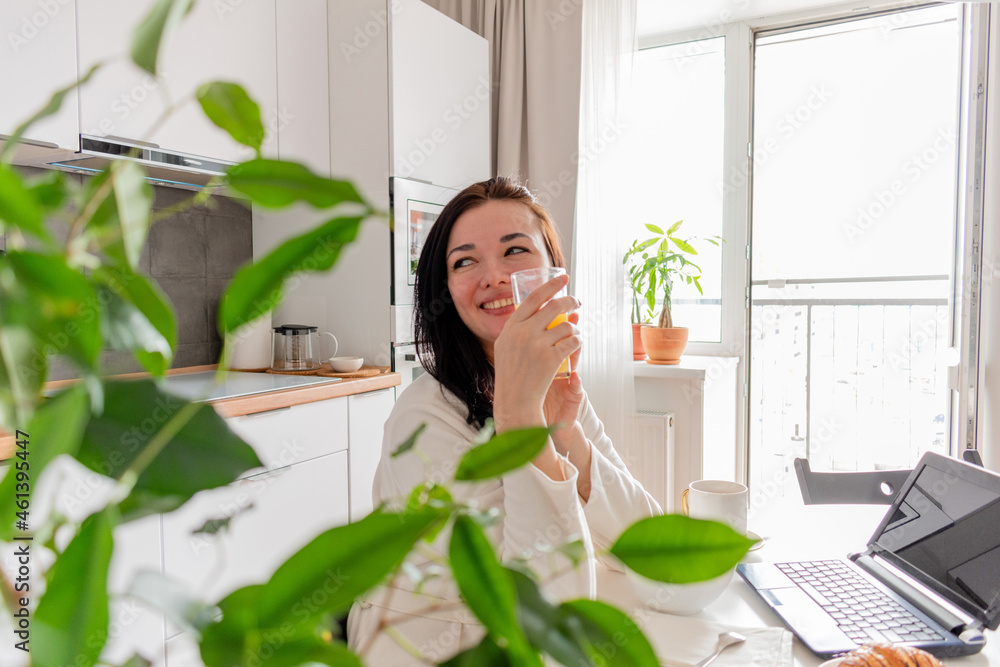 Young woman with black hair in white robe sits on chair the kitchen. Hands hold mug with juice. Young woman with black hair in white robe sits on chair the kitchen. Hands hold mug with juice.