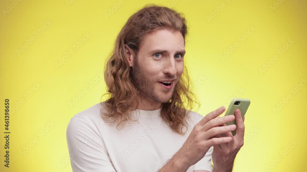 Shocked smiling young Caucasian man in casual whitw T-shirt isolated on yellow background in studio. People lifestyle concept. Boy looks surprised wow hold using mobile cell phone. Shocked smiling young Caucasian man in casual whitw T-shirt isolated on yellow background in studio. People lifestyle concept. Boy looks surprised wow hold using mobile cell phone.