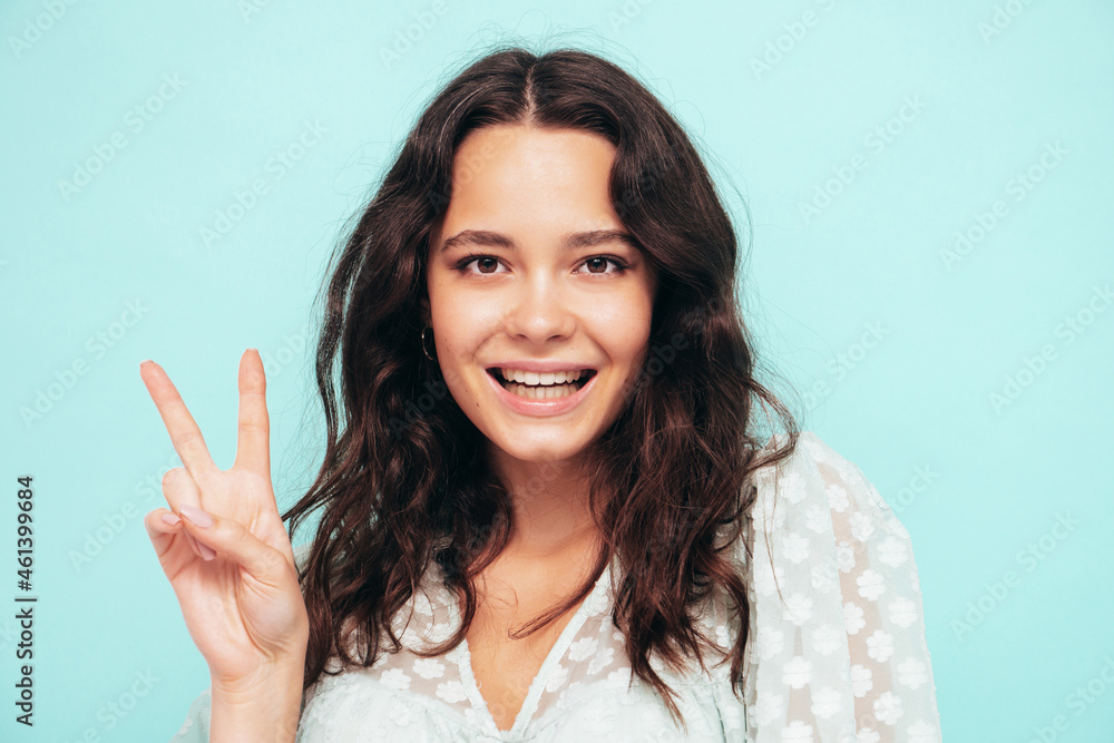 Closeup portrait of young beautiful smiling female in trendy summer dress. Sexy carefree woman posing near blue wall in studio. Positive model having fun indoors. Shows peace sign Closeup portrait of young beautiful smiling female in trendy summer dress. Sexy carefree woman posing near blue wall in studio. Positive model having fun indoors. Shows peace sign
