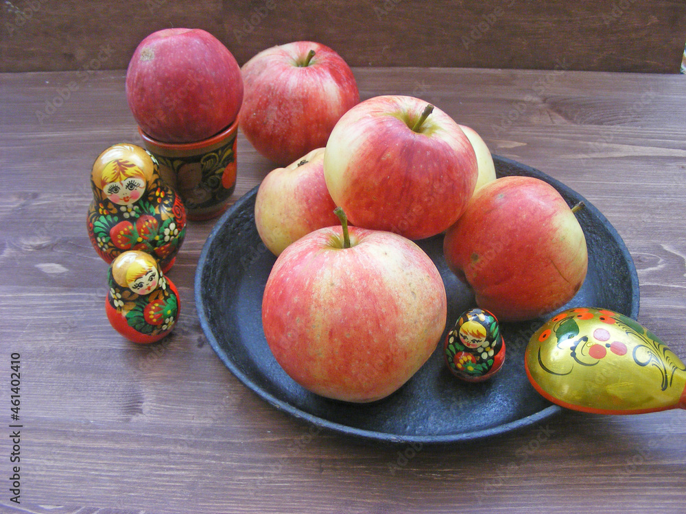 Composition of red and yellow apples on black cast iron plate with traditional russian nesting dolls matrioshka and painted spoon on brown wooden table Composition of red and yellow apples on black cast iron plate with traditional russian nesting dolls matrioshka and painted spoon on brown wooden table
