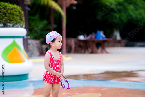 Portrait of Asian baby girl playing water in the water park. Child wearing red swimsuit and pink swim cap. Happy kid smiling.