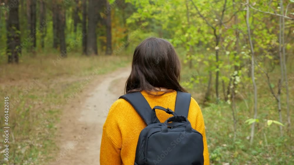 Long haired brunette woman with black backpack wearing orange sweater walks along empty narrow road across green forest backside view Long haired brunette woman with black backpack wearing orange sweater walks along empty narrow road across green forest backside view