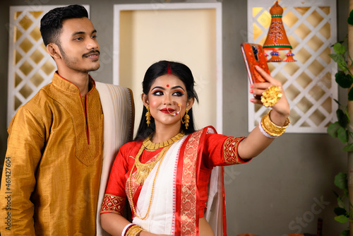 Photography Portrait of Indian man dressed in kurta pajama with beautiful Indian woman wearing traditional Indian saree, gold jewellery and bangles, holding mobile phone taking selfie photo using smartphone