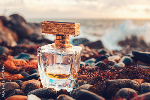 A golden transparent perfume bottle with drops on a wet pebble beach. Close-up. In the background, the sea surf is blurred. Perfume advertising concept photo