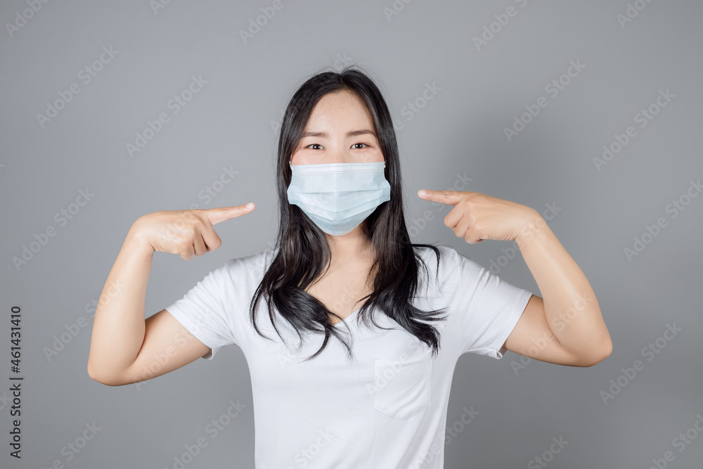 Portrait of Asian woman pointing to a medical face mask to protect Covid-19 (Coronavirus) wears white t-shirt on gray background Portrait of Asian woman pointing to a medical face mask to protect Covid-19 (Coronavirus) wears white t-shirt on gray background