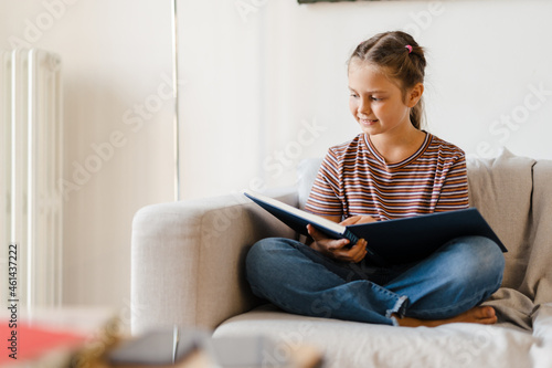 White preteen girl smiling and reading book while sitting on couch