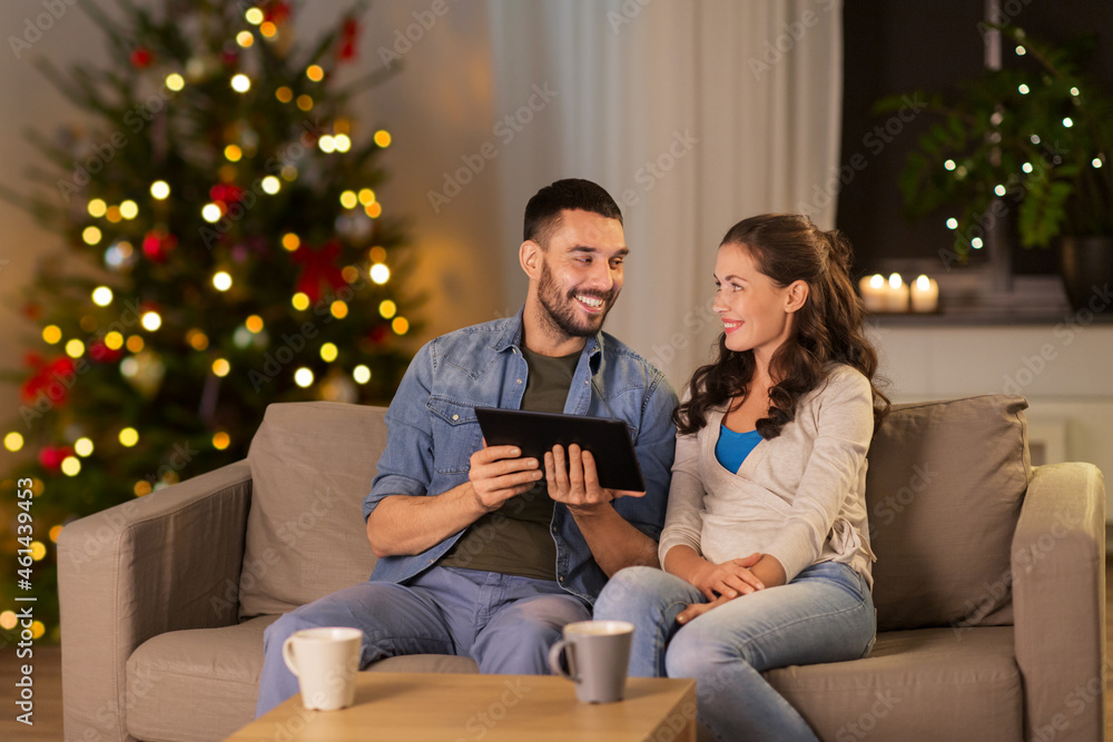 Obraz premium technology, winter holidays and people concept - happy couple using tablet computer at home in evening over christmas tree lights on background