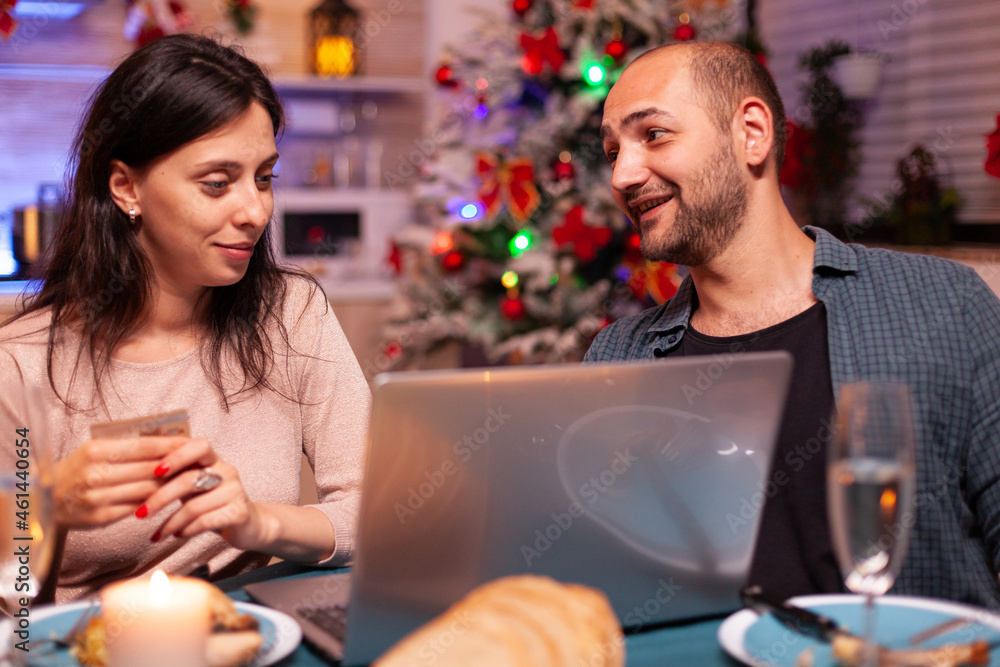 Cheerful couple buying xmas gift present paying online with credit card on laptop computer sitting at dining table in x-mas decorated kitchen. Happy family shopping for winter holidays