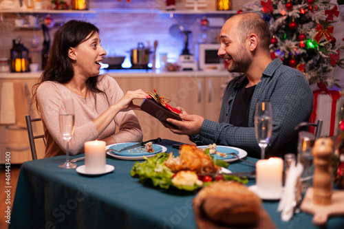 Happy couple spending christmas winter holiday surprising with xmas present with ribbon on it sitting at table in x-mas decorated kitchen. Joyful family celebrating christmastime together