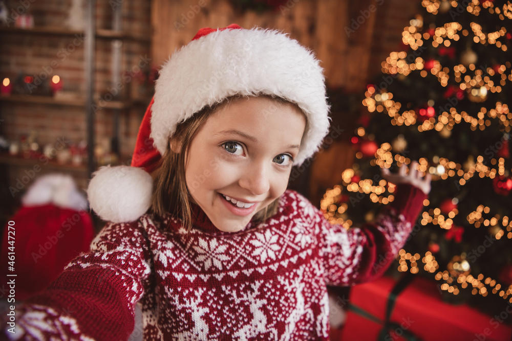 Photo of adorable blogger little kid girl make selfie showing evergreen tree wear ornament pullover santa hat indoors Photo of adorable blogger little kid girl make selfie showing evergreen tree wear ornament pullover santa hat indoors