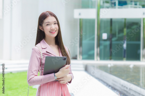 Asian professional business female with long hair is smiling outdoor in the garden front of the office while hold at the tablet in her hand to go out meeting with clients at the office. 