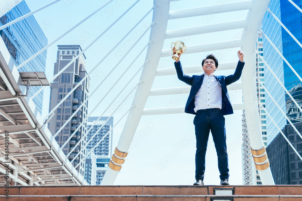 Successful businessman celebrating his victory with arms up and holding gold trophy on cityscape background.