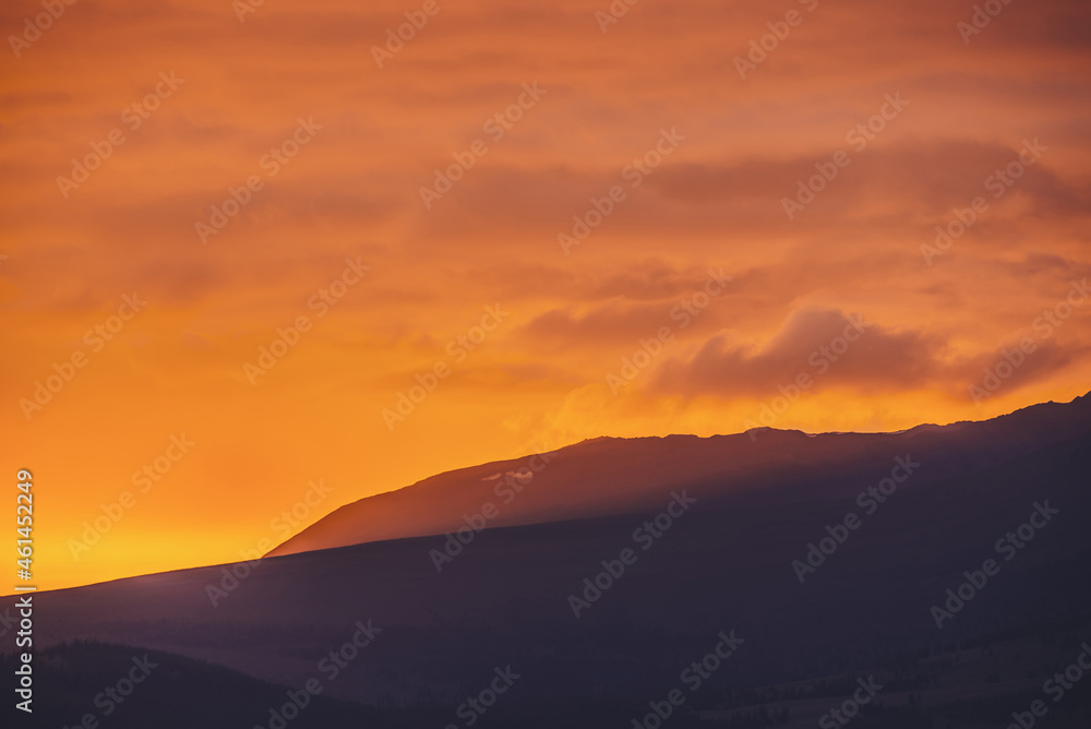 Atmospheric landscape with dark silhouettes of mountains on background of vivid orange dawn sky. Colorful scenery with sunset or sunrise of illuminating color. Sundown paysage with sunbeam on mountain