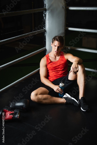 tired man in sportswear holding sports bottle while sitting on floor near boxing gloves.