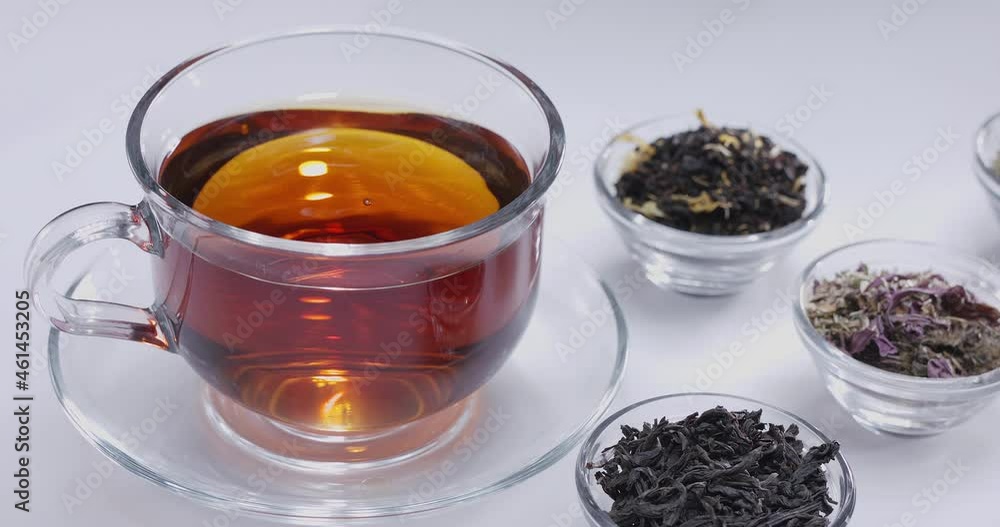 Drop slowly falls into a glass cup of tea. Around there are small bowls with different types of leaf tea. White background. Drop slowly falls into a glass cup of tea. Around there are small bowls with different types of leaf tea. White background.