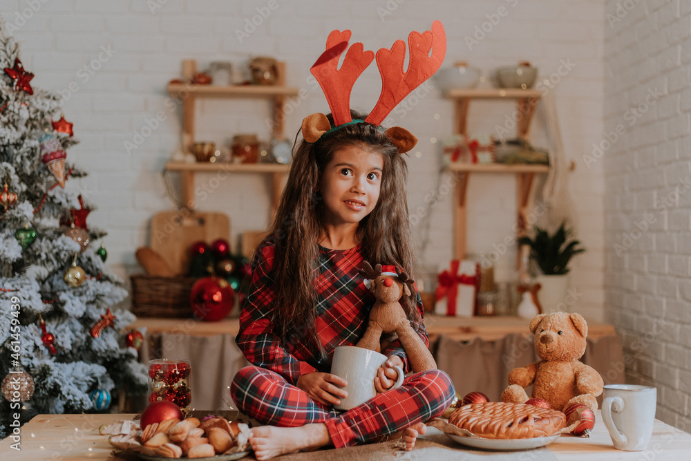 Fototapeta premium cute little brunette girl in checkered red pajamas with reindeer horns on her head is eating a Christmas cake and drinking tea from a white mug in a beautifully decorated kitchen. lifestyle, hygge
