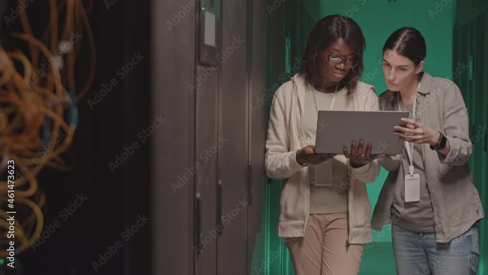 Medium long of intelligent African American woman holding portable computer, her Caucasian female colleague looking at screen, standing in green-lighted server room Medium long of intelligent African American woman holding portable computer, her Caucasian female colleague looking at screen, standing in green-lighted server room
