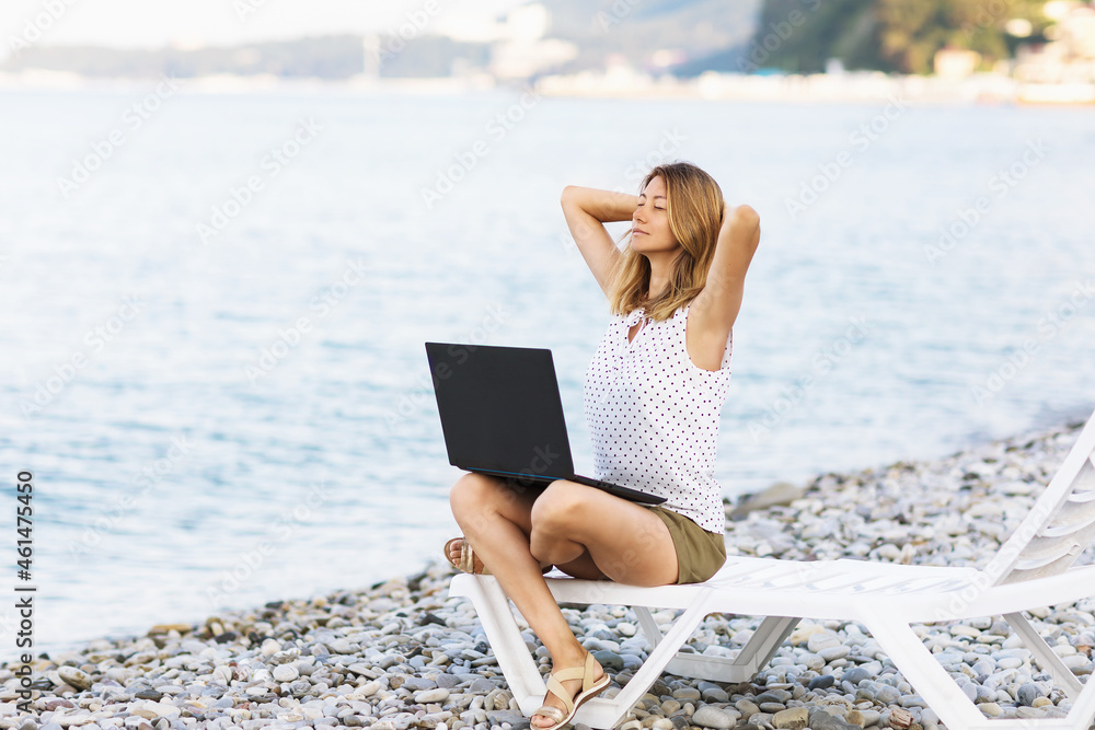 A woman sits on a sun lounger with a laptop on the seashore, combines rest and work