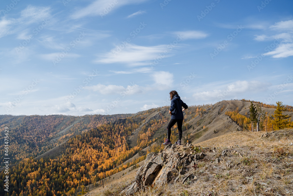 A girl in dark clothes walks along the top of the mountain, looks at the mountains and forest, the clear sky overhead, the autumn landscape.