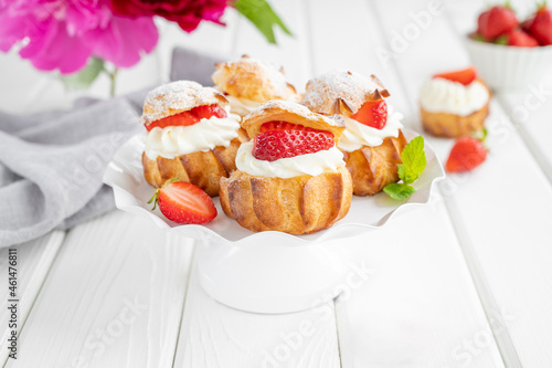 Delicious homemade small cakes profiterole choux pastry with custard, strawberry and icing powder on the white wooden background. Copy space.