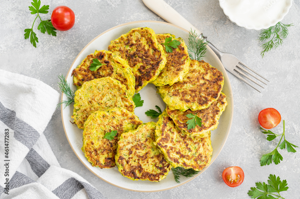 Vegetable zucchini pancakes with cheese, dill and parsley on a plate with sour cream on a gray concrete background. Top view, copy space. Vegetable zucchini pancakes with cheese, dill and parsley on a plate with sour cream on a gray concrete background. Top view, copy space.