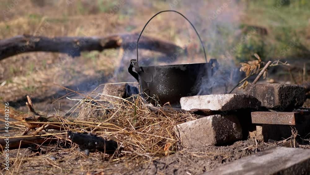 Cooking Food in a Saucepan Over an Open Bonfire in Smoke at Sunset. A black tourist boiler in soot smokes in sunlight on dry grass. Concept of camping, tourist recreation, nature survival. Summertime. Cooking Food in a Saucepan Over an Open Bonfire in Smoke at Sunset. A black tourist boiler in soot smokes in sunlight on dry grass. Concept of camping, tourist recreation, nature survival. Summertime.