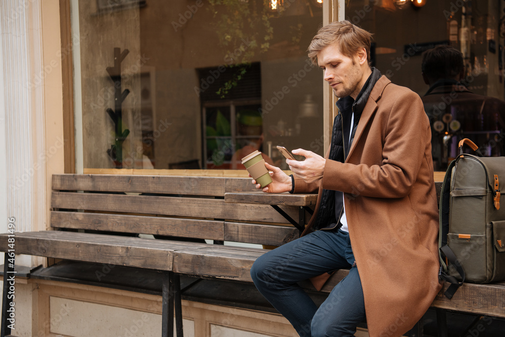 Handsome man holding takeaway coffee cup and mobile phone in hand. City life cafe in background. Man in brown coat Modern businessman with backpack - obrazy, fototapety, plakaty 