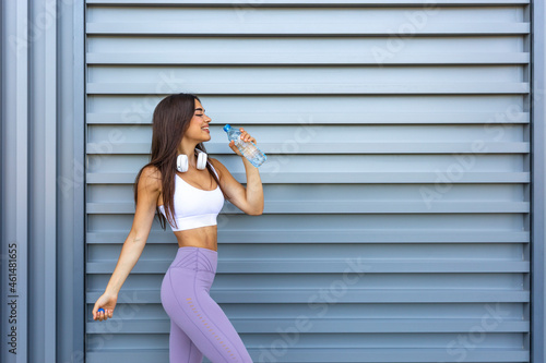 Shot of beautiful female runner standing outdoors holding water bottle. Fitness woman taking a break after running workout. Cheerfully smiling sporty woman drinking water after work out exercising