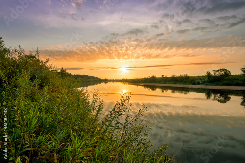 Scenic view at beautiful summer river sunset with reflection on water with green bushes, calm water ,deep colorful cloudy sky and glow on horizon on a background, spring evening landscape